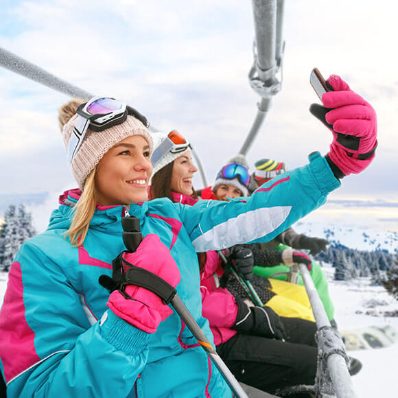 Coton Textilreinigung - Reinigung Skibekleidung - Frau im Sessellift mit anderen Skifahren beim Selfie